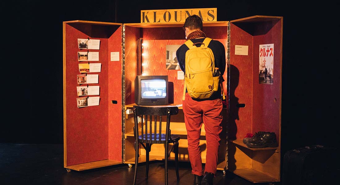 Photo d'une partie de l'exposition. Un jeune homme de dos observe une sorte d'armoire ouverte où s'entrepose à l'intérieur plusieurs éléments de l'exposition : des tableaux, une télévision,une chaise...