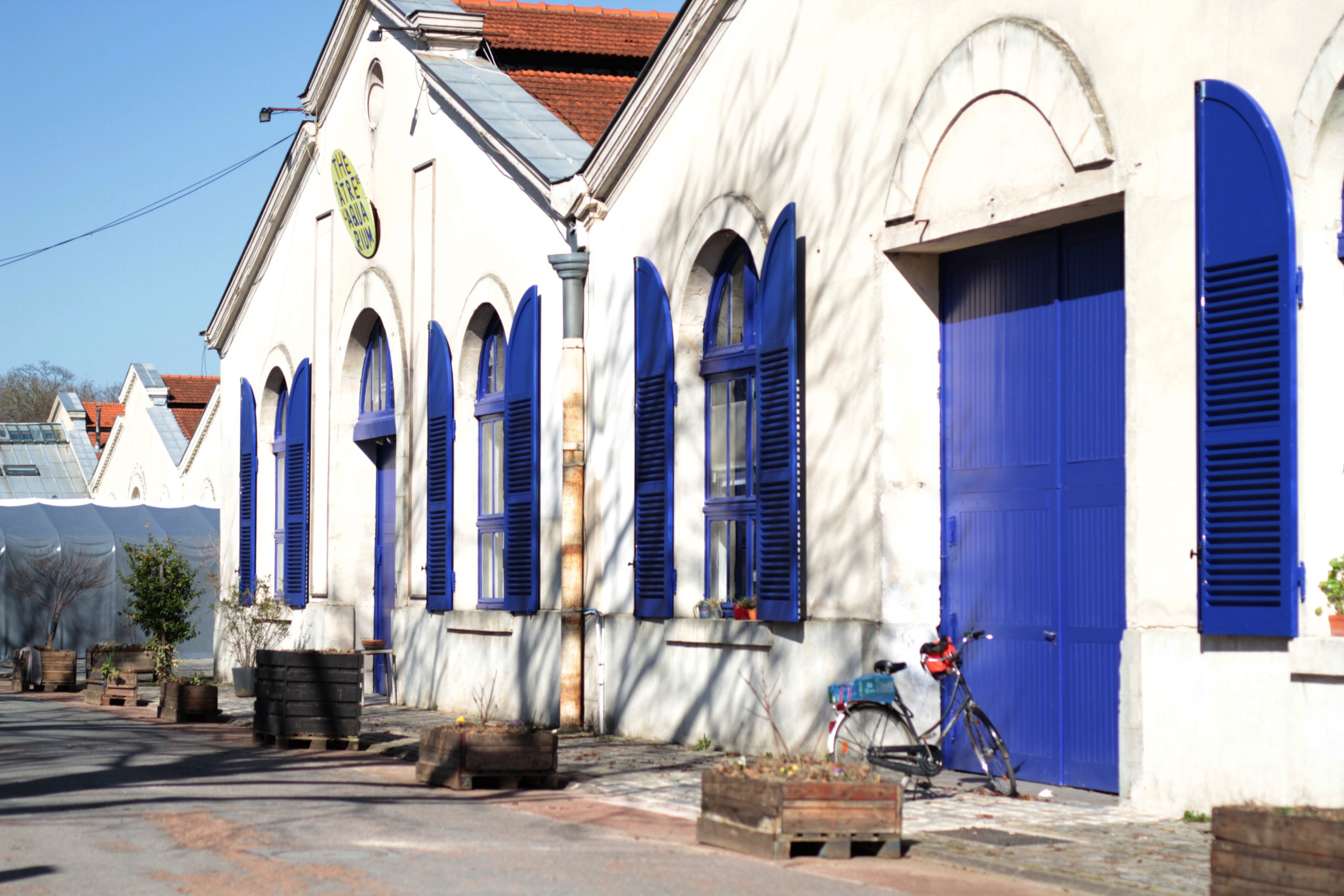 Photo de la façade du théâtre de l’aquarium, prise depuis la route d’accès. Mur en pierre blanc et volets en bois bleus.