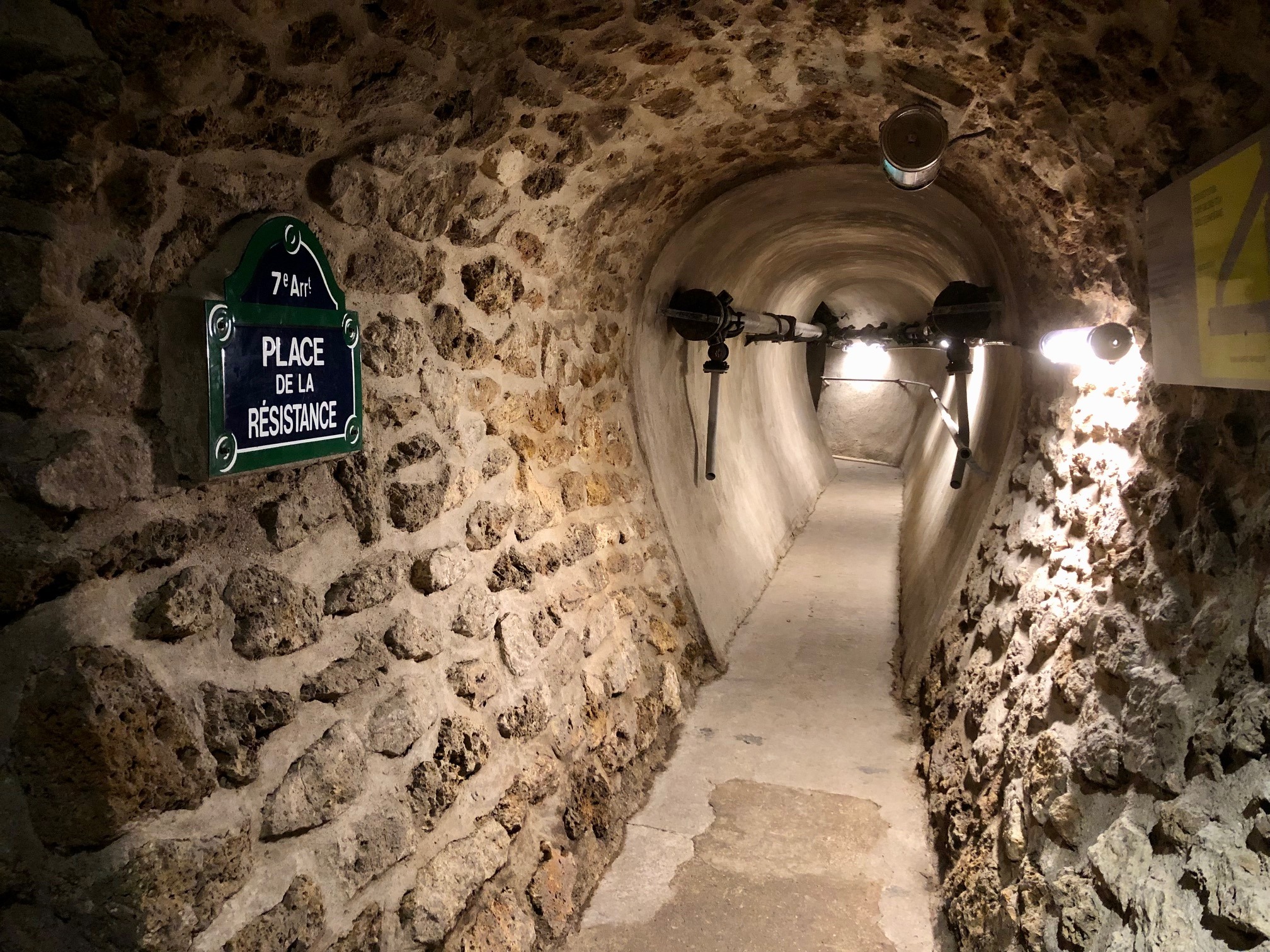 photo couleur d'une allée dans les égouts de Paris. Elle représente une entrée de tunnel en vieilles pierres apparentes, sur le côté gauche de l'entrée, un odonyme (Plaque de rue) bleu et contours vert foncés avec les indicateurs en blanc : septième arrondissement, Place de la Résistance.