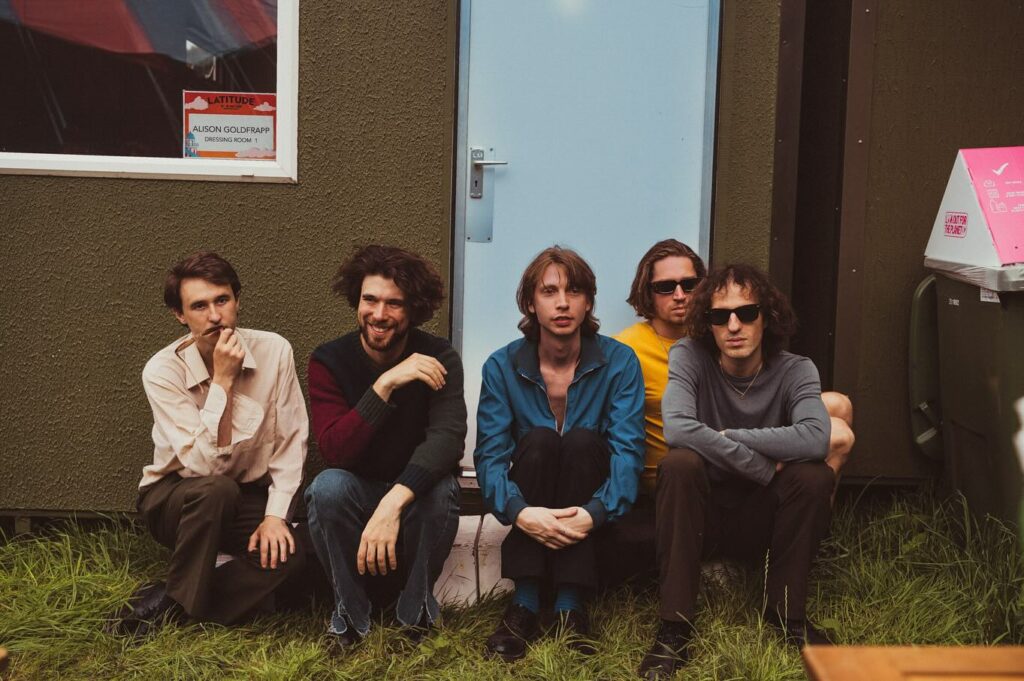 Photo en couleurs du groupe, composé de 5 musiciens. Ils sont assis ôte à côte sur des moellons de béton blanc devant un baraquement, habillé dans un style années 70. Le baraquement est lui aussi empreint aux années 70, les murs sont en crépis marron avec une porte bleu très clair, de l'herbe se trouve au sol. Sur le côté gauche on distingue une vitre. Une affichette est collée, il y est écrit "latitude, Alison Goldfrapp, dressing room 1"