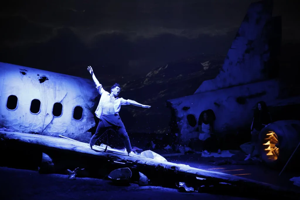 Photo prise lors d'une représentation. C'est la nuit, on distingue en décor un crash d'avion. Celui ci est coupé en deux. La lumière se porte sur un homme qui danse sur l'aile de l'avion. Ses bras et ses jambes sont écartés, son corps forme un X.