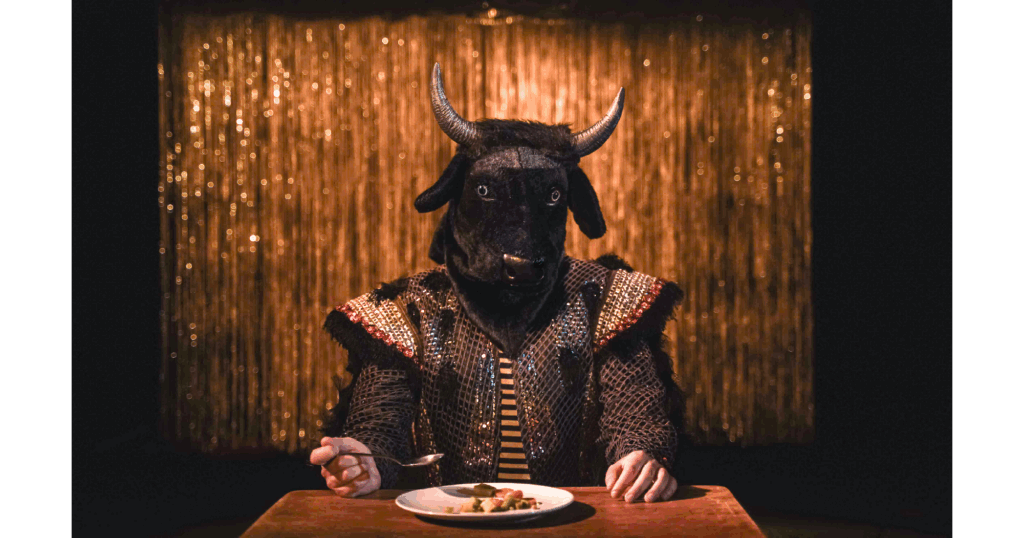 Photographie :Une personne assise à une table, avec une tête de taureau noire et des cornes. Elle tient une cuillère devant une assiette de nourriture. En arrière-plan, un rideau doré scintillant.