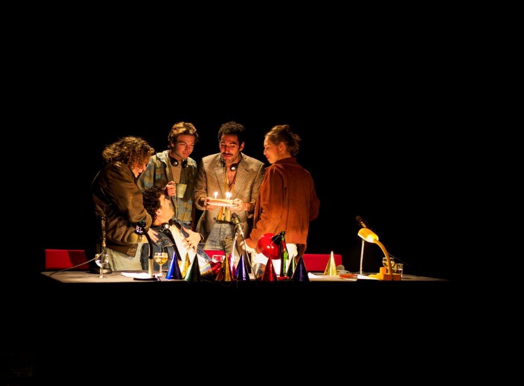 Photographie : Cinq personnes rassemblées autour d’une table éclairée dans une ambiance théâtrale sombre. L’une d’elles tient un petit gâteau avec des bougies allumées. La table est décorée de chapeaux pointus colorés et de lampes de bureau. Les personnages semblent célébrer un moment festif sur scène.