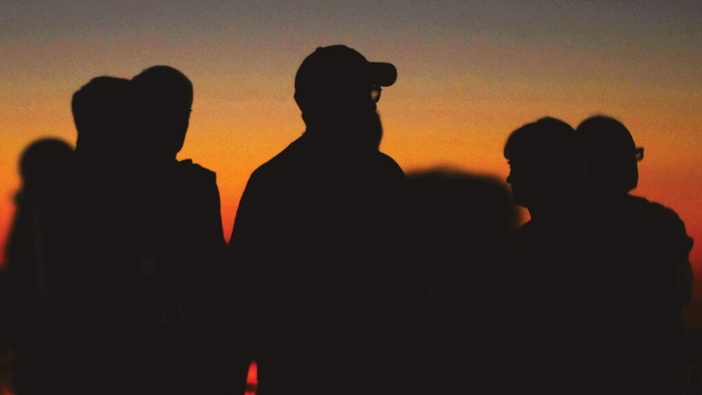 Photographie : Silhouettes de plusieurs personnes vues de profil et de dos, se détachant dans l’obscurité devant un ciel au coucher du soleil aux teintes orange.