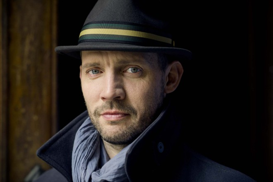 Photographie : Portrait d’un homme portant un chapeau noir avec une garniture à bandes jaune et verte, un manteau foncé au col relevé et une écharpe grise. Il regarde l’objectif avec une expression neutre, sur un fond sombre qui met son visage en valeur.