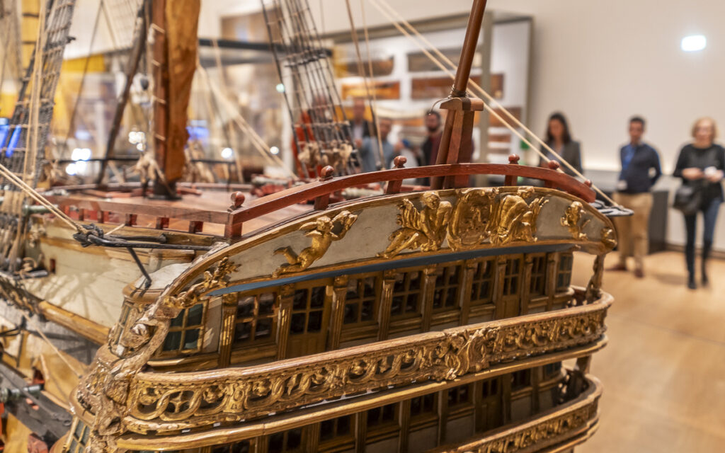 photographie : Maquette détaillée d’un grand navire ancien exposée dans un musée, avec plusieurs visiteurs debout en arrière-plan.