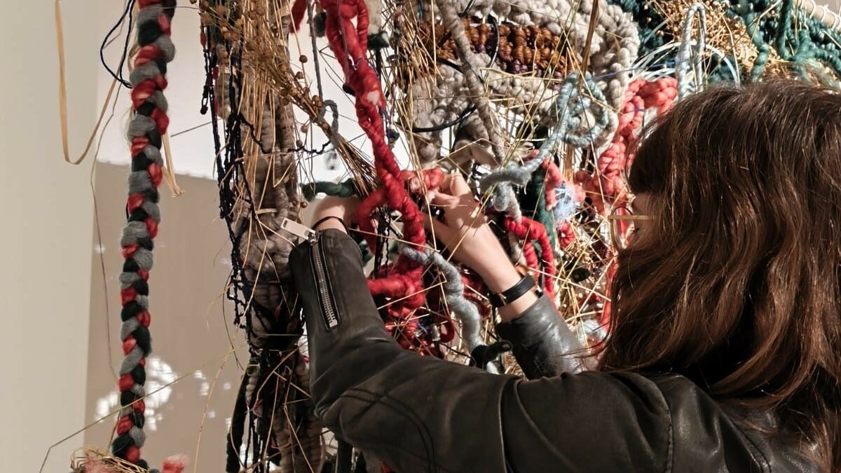 Photographie, une personne de dos, portant une veste en cuir, en train de toucher une grande installation textile composée de cordes épaisses, de fibres colorées.