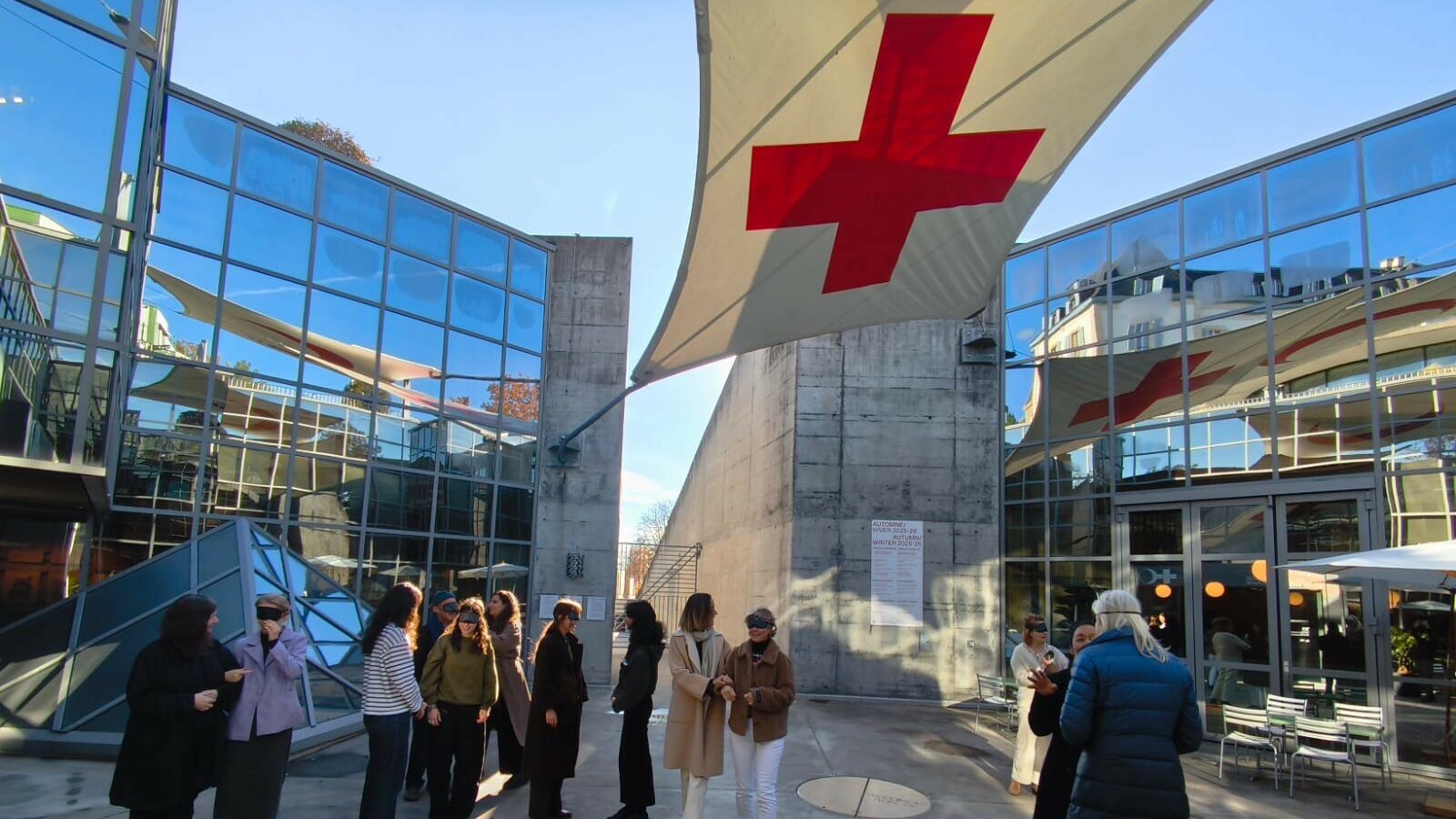 Photographie : plusieurs personnes debout dans une cour en plein air, entourée de bâtiments aux façades vitrées, avec une grande toile blanche marquée d’une croix rouge au-dessus d’elles.