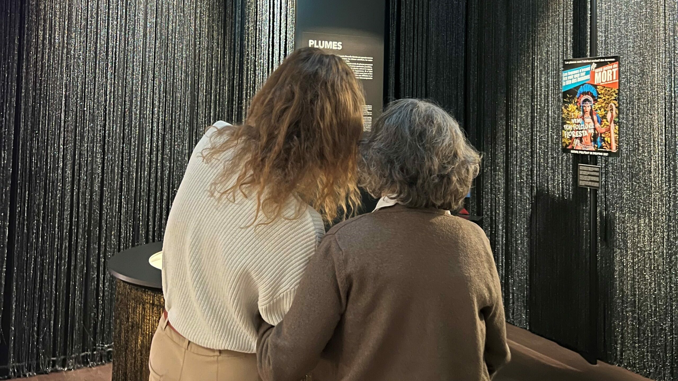 Photographie : Deux personnes debout de dos, observant un panneau d’exposition dans une salle sombre aux murs couverts de rideaux scintillants.