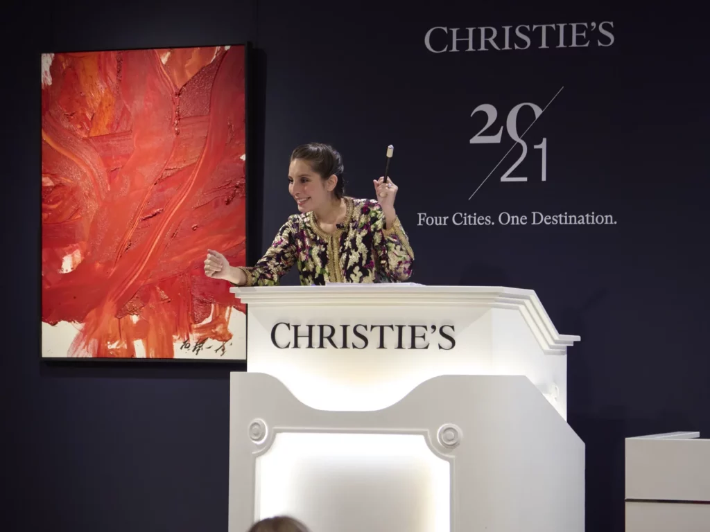 photographie : Commissaire-priseuse dirigeant une vente aux enchères depuis un pupitre Christie's, avec un grand tableau abstrait rouge accroché au mur derrière elle.