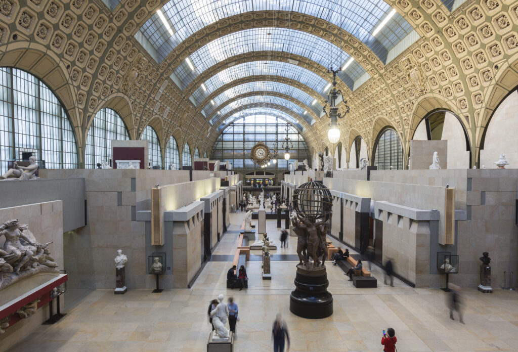 photographie : Grande nef du musée d’Orsay, avec sa verrière arquée et ses sculptures disposées le long de l’allée centrale, où circulent plusieurs visiteurs.