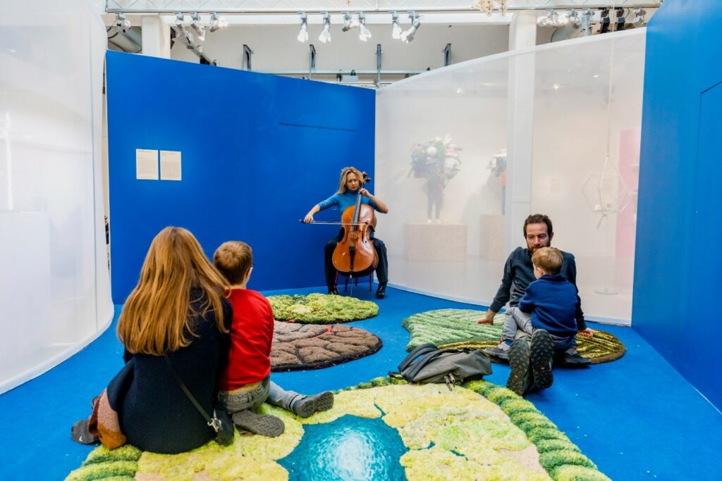 photographie : Musicienne jouant du violoncelle dans une salle d’exposition aux murs bleus, tandis que des adultes et des enfants sont assis au sol sur des tapis colorés pour l’écouter.