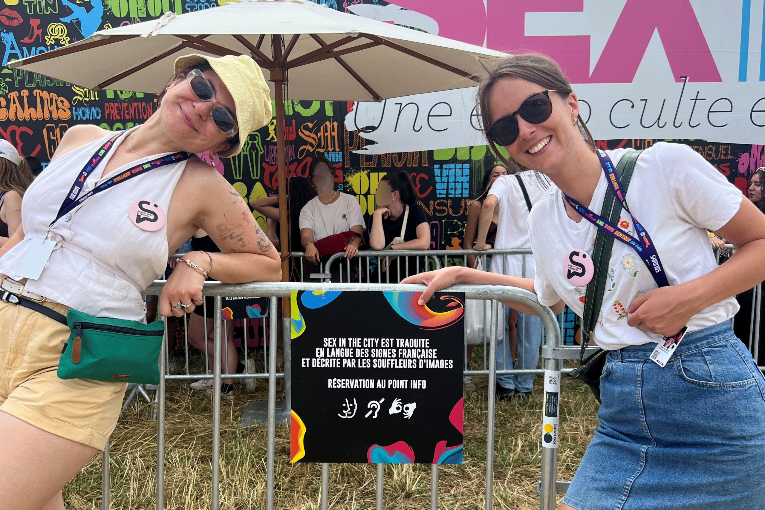 Photographie format paysage : deux femmes posant devant des barrières   où est accroché un panneau. 

Texte du panneau : Sex in the city est traduite en langue des signes française et de décrite par les Souffleurs d'Images.