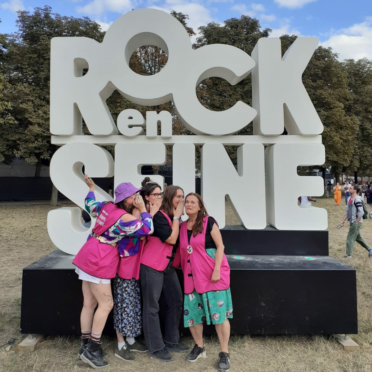 Photographie au format carré : quatre femmes se tiennent côte à côte. Trois d’entre elles placent leur main près de l’oreille de la quatrième, imitant le geste du soufflage. En arrière-plan, une grande installation du logo du festival Rock en Seine.